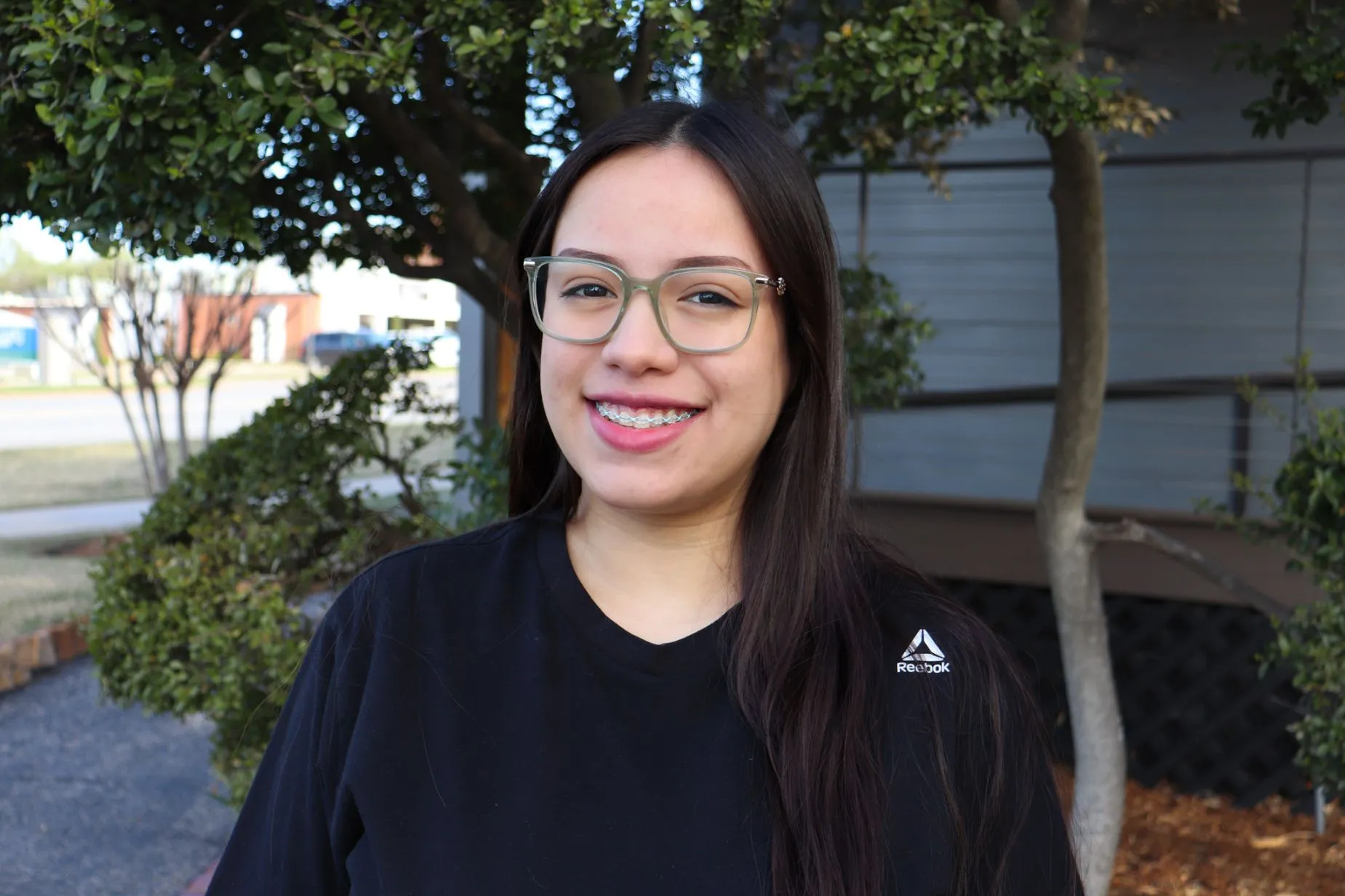Adolescente sonriendo con frenillos y gafas, en un entorno exterior con รกrboles, representando la importancia de la ortodoncia y la confianza en la sonrisa.