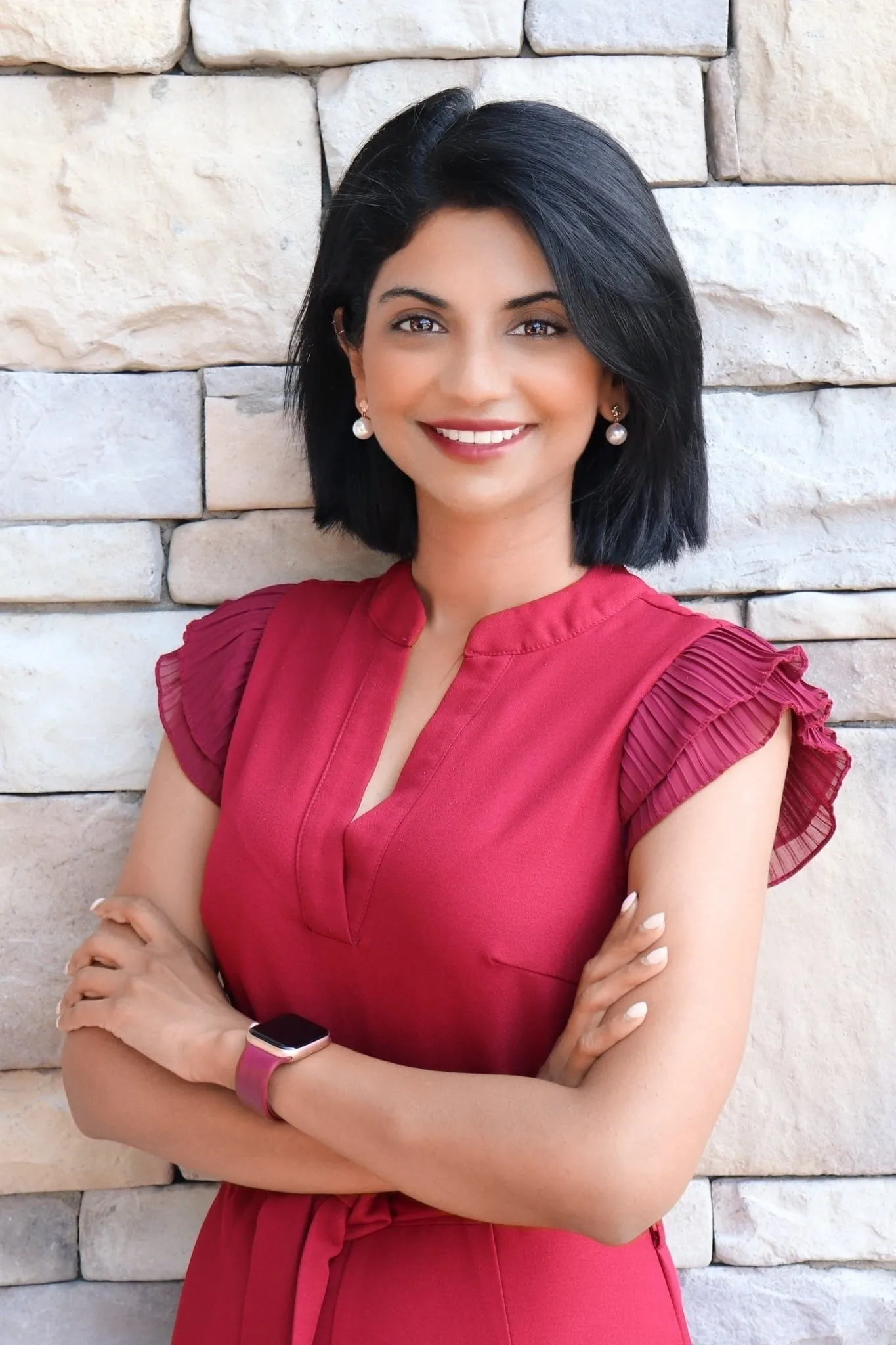 Dra. Pheba Abraham, ortodoncista, sonriendo con brazos cruzados frente a una pared de piedra, vestida con blusa roja, destacando su profesionalismo y calidez en el ámbito de la atención odontológica.